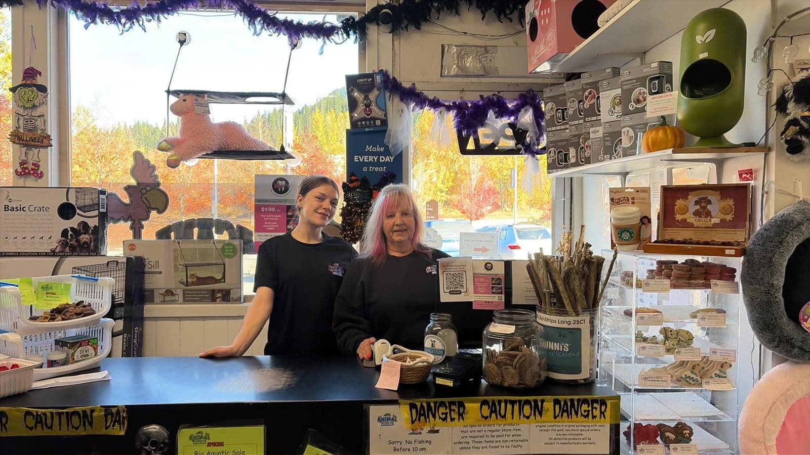 Two smiling staff behind the front counter of the Revelstoke store, with autumn trees through the window behind them